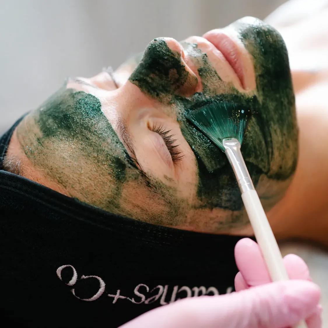 Esthetician applying a KRX Green Sea Peel to a client's face with a fan brush at Peaches + Co